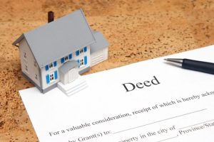 A blue and white model house sits next to a property deed, possibly a lady bird deed, with a pen on top of it.
