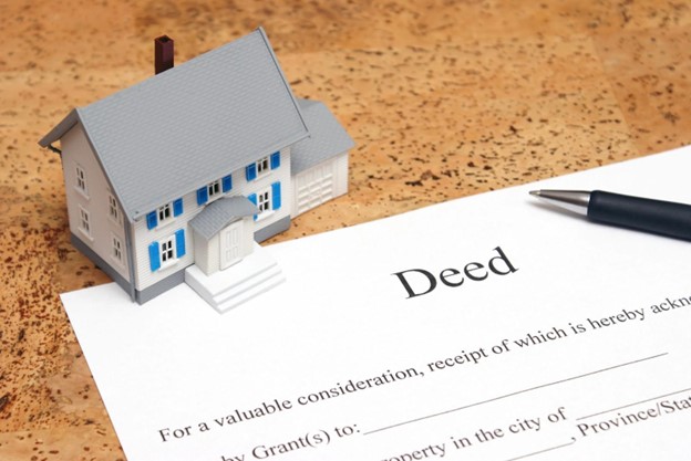 A blue and white model house sits next to a property deed, possibly a lady bird deed, with a pen on top of it.