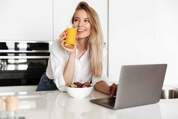 Newly single young woman standing at kitchen island with laptop computer and glass of orange juice; estate planning updates after divorce.