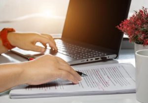 Close-up of a woman’s hands poised over a printed document on the right and a laptop keyboard on the left; preparing a valid Will.