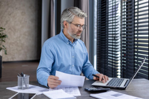 The personal representative of a Florida estate reviews documents related to probate administration, working with hard copies, a calculator, and a laptop computer at a table next to blinds letting in natural light.